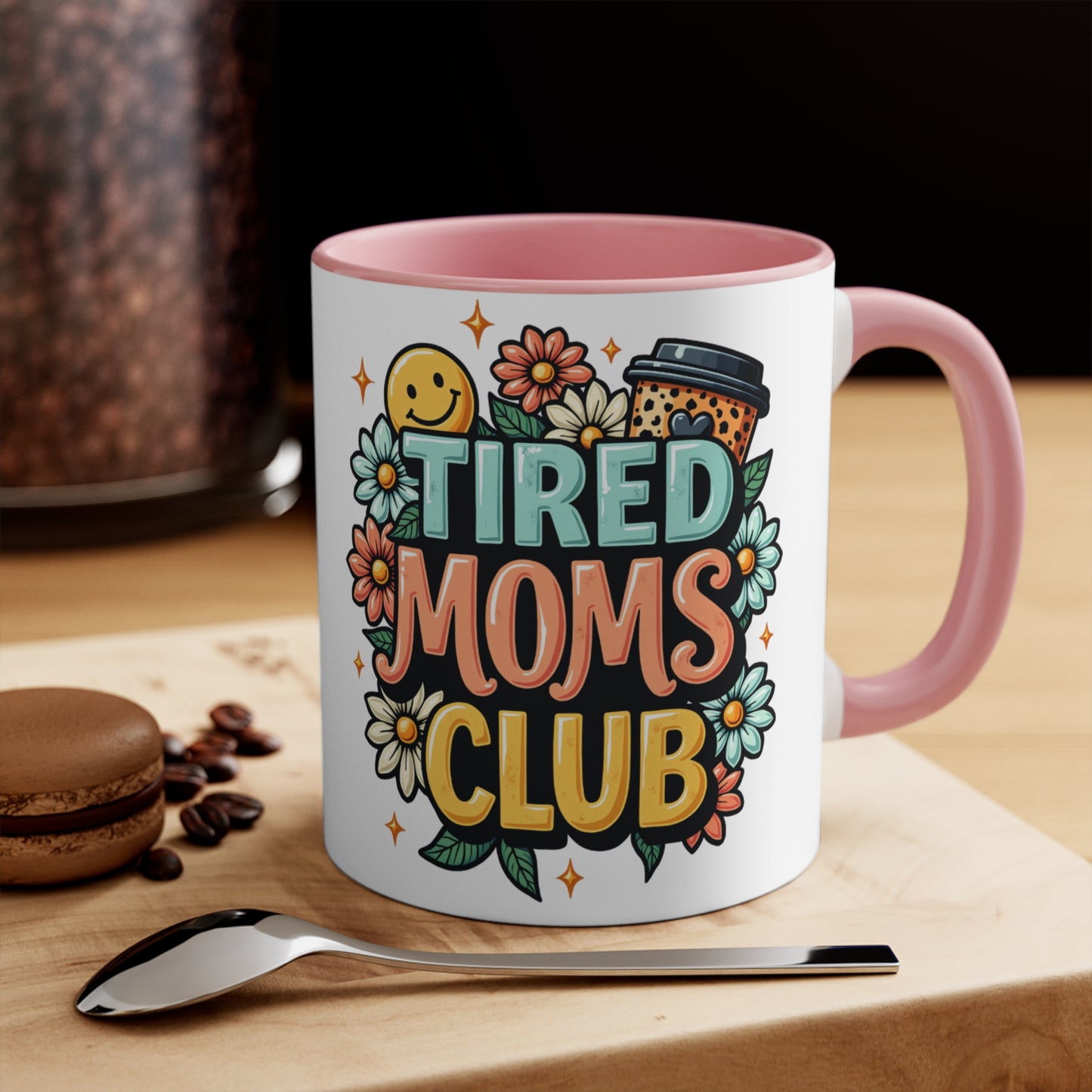 White mug with pink handle, "Tired Moms Club" text, flowers, and coffee cup design.