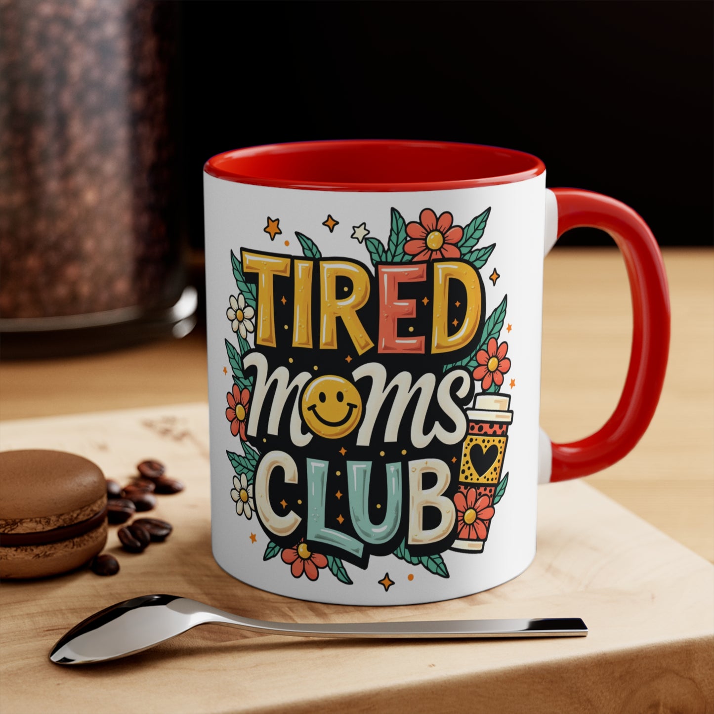 White mug with red handle and "TIRED MOMS CLUB" text and floral designs.