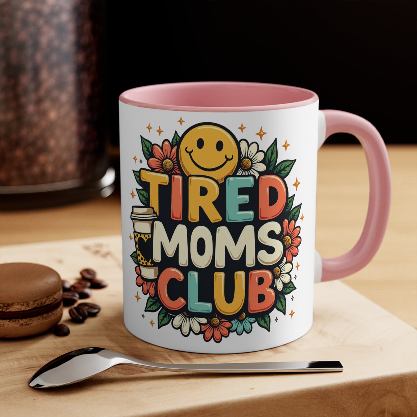 Mug with "Tired Moms Club" text and smiley face, coffee beans, and a macaron.
